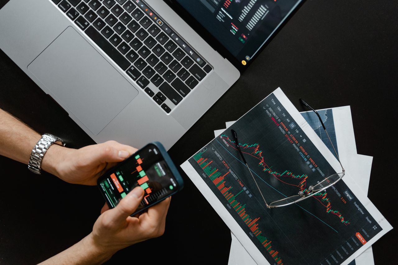home-services-02 Hands using smartphone beside laptop with stock charts, showcasing digital trading.